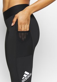 Czarne legginsy sportowe z wysokim stanem, teksturowaną kieszenią boczną i białym logo Adidas w dolnym lewym rogu. Wykonane z gładkiego, elastycznego materiału.