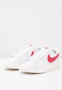 Vita sneakers med röd Nike-swoosh, snörning, slätt lädermaterial, rund tå och texturerad gummisula.