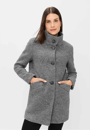Woman wearing a mid-length buttoned gray wool coat with a high collar and hands in pockets, standing against a plain light background.