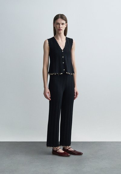 Γυναίκα που φοράει μαύρο αμάνικο τοπ με κουμπιά και ταιριαστά πλεκτά παντελόνια με διακοσμητικό τελείωμα, στέκεται σε γκρίζο πάτωμα μπροστά σε ανοιχτό γκρι τοίχο.