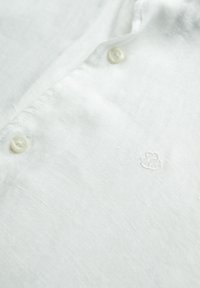 Biała lniana koszula z teksturowanym splotem, dwoma guzikami oraz subtelnym haftowanym logo w jasnej nici. Gładki materiał z luźnym wykończeniem.