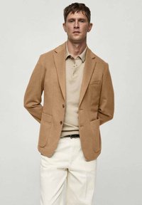 Smeđi blazer od teksturirane tkanine, s dva prednja džepa, prorezanim reverima i dva dugmeta. Nosite ga preko bež polo majice i bijelih hlača.