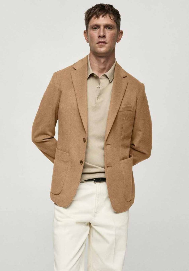 Smeđi blazer od teksturirane tkanine, s dva prednja džepa, prorezanim reverima i dva dugmeta. Nosite ga preko bež polo majice i bijelih hlača.