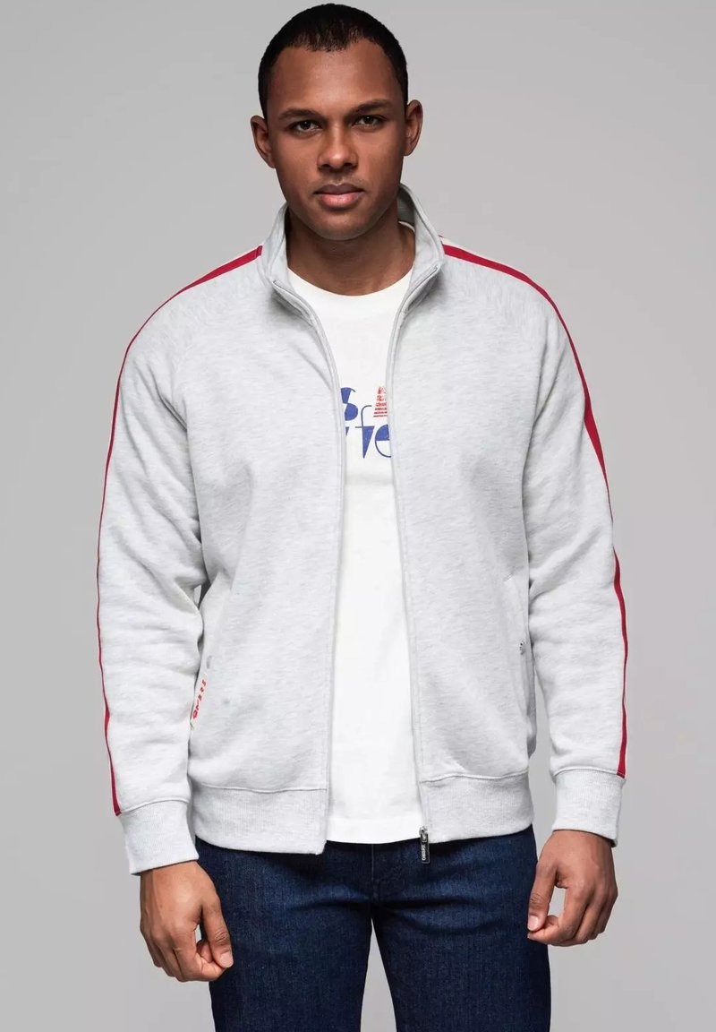Felpa grigia con zip e strisce rosse sui lati, dotata di un collo alto, polsini a costine e una t-shirt bianca sotto. Fit casual, texture liscia.