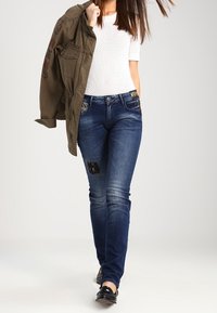 Jean en denim bleu foncé avec des patches déchirés, assorti d'un haut blanc texturé et d'une veste vert olive, avec des détails brodés.