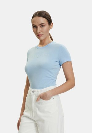 Giovane donna con capelli castani lisci legati in una coda di cavallo indossa un top azzurro a maniche corte e pantaloni bianchi a vita alta, mano in tasca.