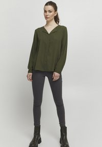 Olive green long-sleeve blouse with a v-neck collar and a front placket. Paired with grey fitted jeans and black ankle boots. Smooth fabric texture.