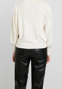 Maglione color crema con un design texturizzato che presenta dettagli in perle disposti in un motivo a rombi sulla schiena, abbinato a pantaloni in pelle neri.