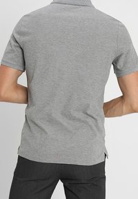 Polo gris avec col, manches courtes et fentes sur les côtés, présenté de dos. Texture de tissu lisse, design simple.