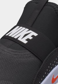 Μαύρο αθλητικό παπούτσι Nike με λευκό λογότυπο στη λουρά και μικρό πορτοκαλί σβούρο στο πλάι της σόλας.