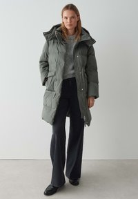 Manteau matelassé gris avec capuche, fermeture éclair et poches latérales. Associé à un pull gris et un pantalon noir à jambes larges. Texture lisse et rembourrée.