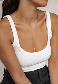 Gouden ketting met een hangende parel hanger, gedragen met een witte geribbelde tanktop. De huidskleur is warm en de tanktop heeft brede bandjes.