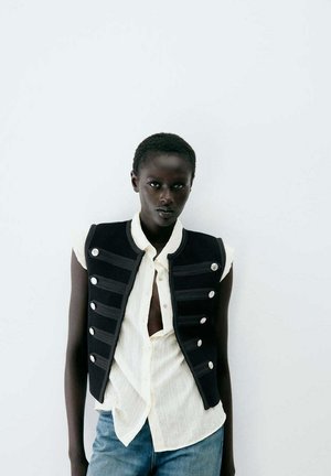 Persona negra con el cabello corto lleva una camisa blanca sin mangas con botones, chaleco negro con botones plateados y vaqueros azules sobre un fondo blanco simple.