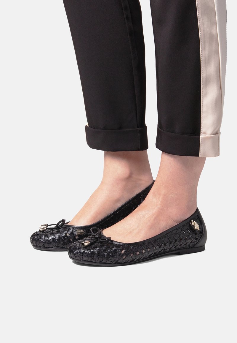 Bailarinas negras tejidas con un acabado brillante, con un lazo decorativo y herrajes metálicos, llevadas con pantalones negros remangados en los puños.