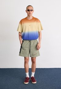 Nike gradient t-shirt su oranžiniu-iki-mėlynu ombre, alyvuogių žalios spalvos šortai su šoninėmis kišenėmis ir taškuota logotipu, baltos kojinės, raudoni sportbačiai.