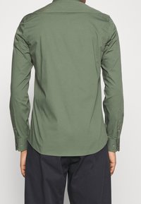 Persoon in een olijfgroen overhemd met lange mouwen en donkere broek, van achteren gefotografeerd tegen een effen lichte achtergrond.
