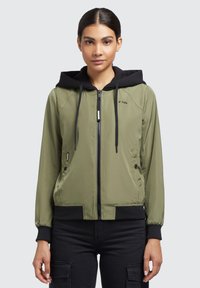 Jachetă bomber verde măslin cu glugă neagră, închidere cu fermoar, buzunare laterale și manșete negre din tricot. Textură netedă cu un design sportiv.