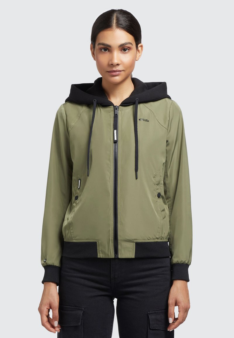 Jachetă bomber verde măslin cu glugă neagră, închidere cu fermoar, buzunare laterale și manșete negre din tricot. Textură netedă cu un design sportiv.
