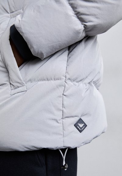 Jachetă căptușită de culoare gri deschis, cu un buzunar cu fermoar, manșete ribbate și un patch cu logo negru pe lateral, având un șnur reglabil la margine.