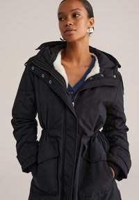 Zwarte parka met een gevoerde capuchon van bont, een getailleerde taille en grote voorkruisen. Voorzien van een rits en knopen als accenten.