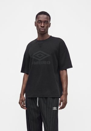 Νεαρός άνδρας που φοράει μια μαύρη μπλούζα Umbro μεγάλης διάστασης και μαύρο παντελόνι με ριγέ υφή και λογότυπο Umbro, στέκεται μπροστά σε απλό λευκό φόντο.