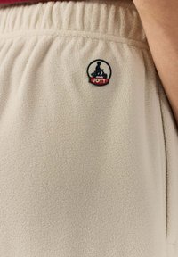 Beige fleece joggers met een elastische tailleband en een JOTT logo patch met daarop een persoon op een scooter. De textuur lijkt zacht en gezellig.