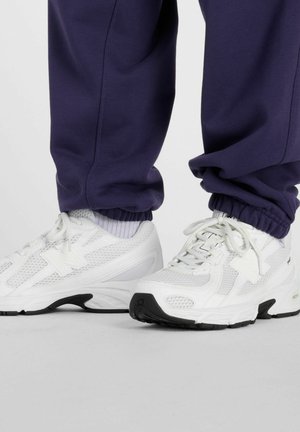 Person iført hvide atletiksneakers med sorte såler og marineblå joggebukser, stående på en grå overflade.