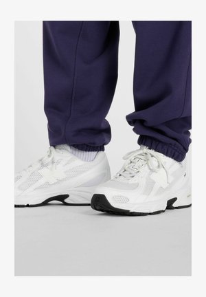 Person iført hvide atletiksneakers med sorte såler og marineblå joggebukser, stående på en grå overflade.