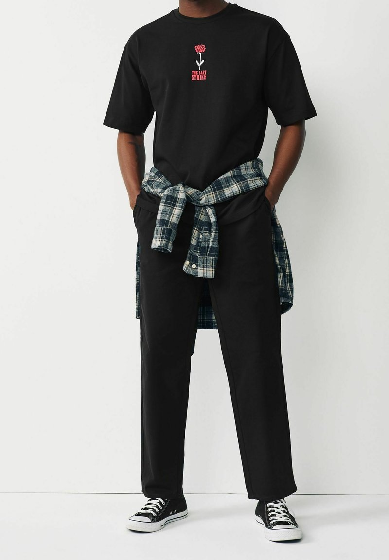 Personne portant un t-shirt noir avec une rose rouge et un texte, un pantalon noir, des baskets noires et blanches, et une chemise à carreaux nouée autour de la taille. Mains dans les poches.