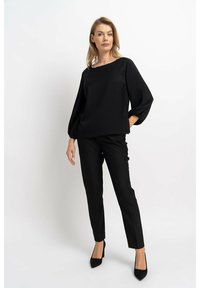 Schwarze Bluse mit Puffärmeln, runder Ausschnitt verziert mit dezenten Verzierungen, kombiniert mit engen schwarzen Hosen und hohen Schuhen.