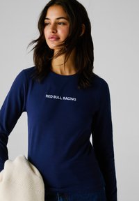 Ženska s temnimi lasmi nosi temnomodro majico z dolgimi rokavi z napisom "RED BULL RACING" ter v eni roki drži zložen bež pleten pulover.