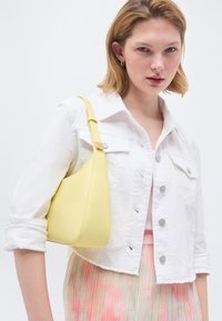 Νεαρή γυναίκα που φοράει λευκό cropped τζιν μπουφάν, πλισέ παστέλ φούστα και κρατάει κίτρινη τσάντα ώμου στον δεξί της ώμο.