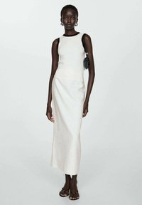 White sleeveless top with dark straps, paired with a long, light-colored skirt. Accessories include a small black bag and minimalist sandals.