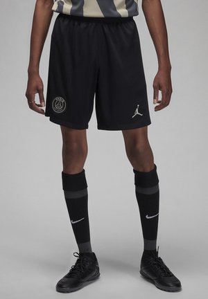 Sportowiec ubrany w czarne sportowe spodenki Paris Saint-Germain z logo Jumpman, czarne skarpetki Nike i czarne buty sportowe na szarym tle.