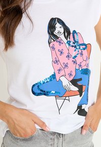 Camiseta blanca con un diseño gráfico colorido de una figura sentada con acentos de lentejuelas en azul y rosa, resaltando patrones florales.