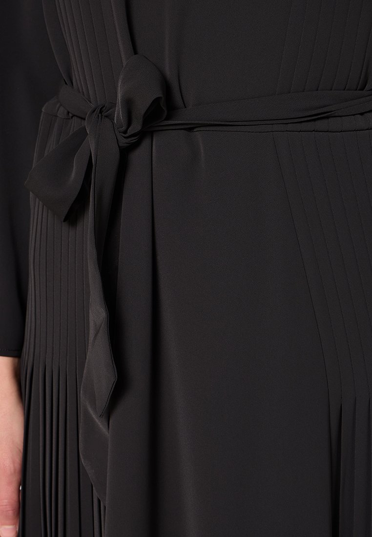 Abito nero con lati plissettati e una cintura di satin annodata a fiocco in vita, che mostra parzialmente una mano e un braccio sinistro.
