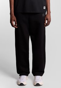 Schwarze Jogginghose mit Logoaufnäher; lockere Passform und weiche Textur. Kombiniert mit hellen Sneakern und einem schwarzen Shirt.