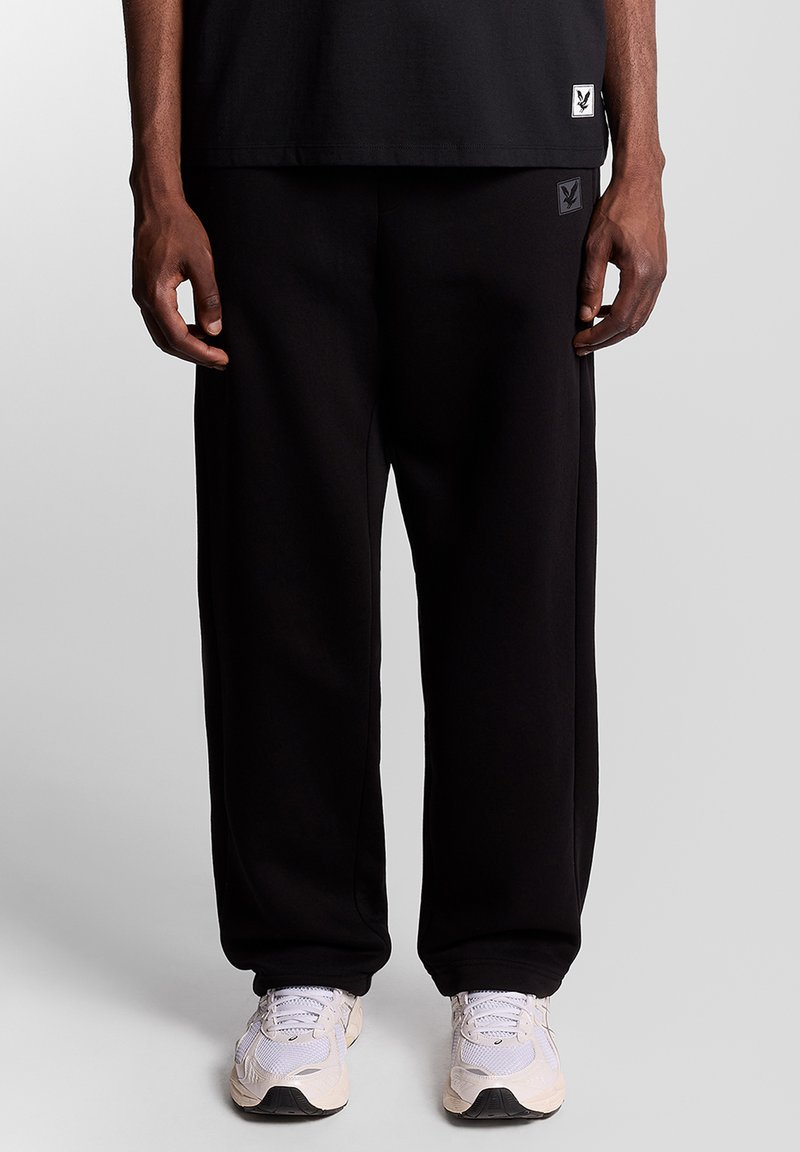 Schwarze Jogginghose mit Logoaufnäher; lockere Passform und weiche Textur. Kombiniert mit hellen Sneakern und einem schwarzen Shirt.