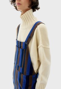 Maglione in maglia avorio con alto collo a costine abbinato a una salopette a righe marrone e blu, con bottoni e una tasca anteriore.