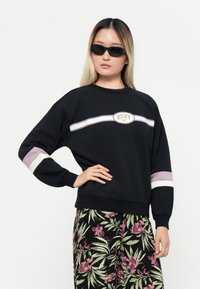 Sudadera negra con el logotipo de Roxy; presenta rayas moradas y blancas en las mangas. El modelo lleva gafas de sol negras y pantalones con estampado floral.