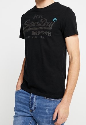 Schwarzes kurzärmeliges T-Shirt mit einem markanten "SuperDry"-Logo in dunklem Grau und blauen Akzenten, aus weichem Baumwollmaterial gefertigt.