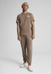 Brun bomulds t-shirt og matchende bukser med Puma-logo. Glat tekstur, afslappet pasform. Modellen bærer hvide sneakers med røde accenter.