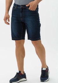 Tummansiniset farkkuhousut suoran leikkauksen kanssa, joissa on viisi taskua ja kontrastiväriset tikkaukset. Yhdistetty urheilullisiin tennareihin.