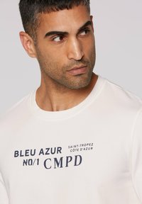 White crew-neck t-shirt featuring navy blue text that reads "BLEU AZUR NO/1 CMPD SAINT-TROPEZ CÔTE D'AZUR." Smooth fabric texture.