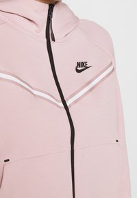 Felpa Nike rosa chiaro con logo nero, chiusura a zip e distintive strisce orizzontali bianche sul petto. Tessuto morbido.