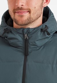 Dark green padded jacket with a high collar, black zipper, adjustable drawstrings, and a smooth, textured finish.