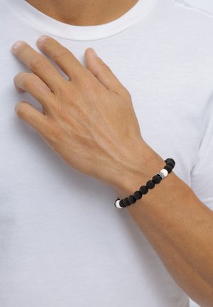 Schwarzes Lavastein-Armband mit weißen Akzentperlen, das eine glatte Textur und ein dehnbares Design aufweist, getragen an einer Hand vor einem hellen Hintergrund.