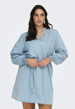 Kvinde iført en løs, lyseblå denimkjole med rynkede ærmekanter, der holder i taljebåndet, og med blå ovalformede øreringe og ringe.