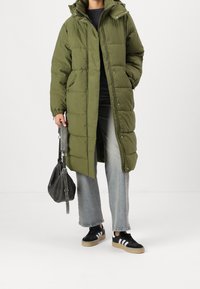 Olive green puffer coat with a hood, snap buttons, and elastic cuffs. Paired with light blue jeans and black sneakers with white stripes.