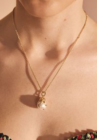 Zelta ķēdes kaklarota ar lielu pērļu piekariņu, kam ir fakturēts zelta vāciņš un nelieli akcentējoši elementi. Gluda, mirdzoša virsma.